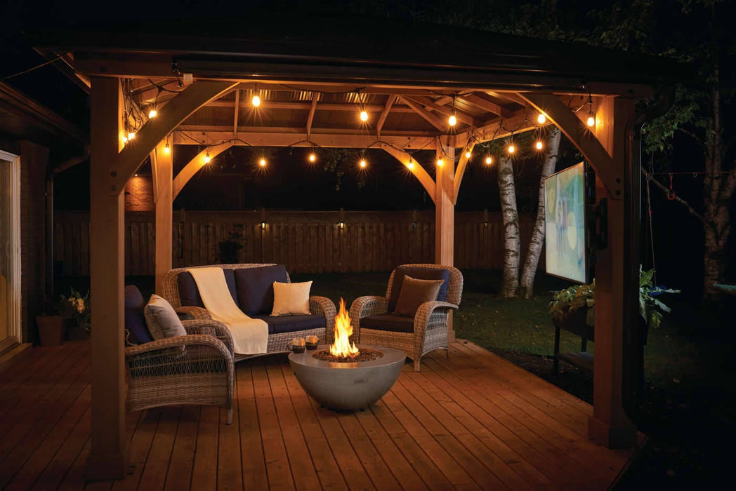 Outdoor patio setup with a fire bowl centerpiece, cozy seating under a lit wooden pergola, and a backyard view at night.