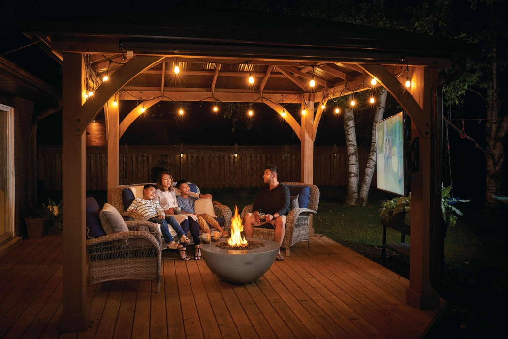 Group of people enjoying an evening around a fire bowl under a lit wooden pergola, with comfortable seating on a backyard patio.