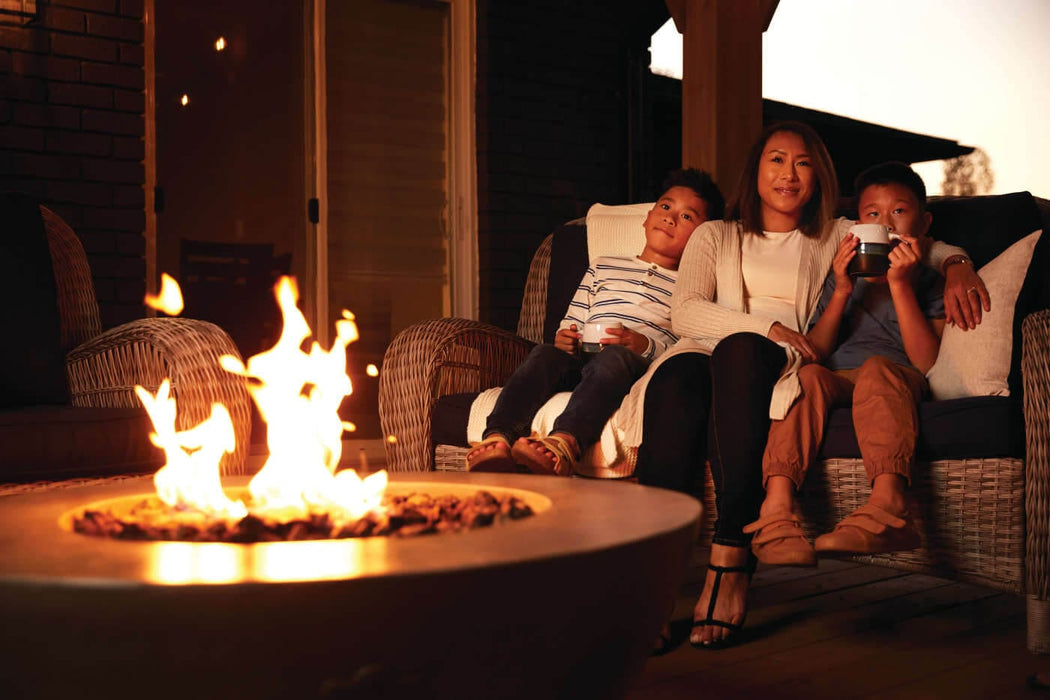 Family sitting on an outdoor wicker couch, enjoying a cozy fire pit in the foreground, with warm ambient lighting on a patio.