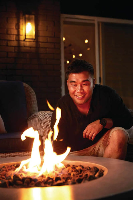 Man crouching near a fire pit with flames, enjoying the warmth, set against a cozy outdoor seating area with soft lighting.