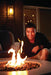 Man crouching near a fire pit with flames, enjoying the warmth, set against a cozy outdoor seating area with soft lighting.