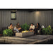 Outdoor patio scene with people sitting on wicker furniture around a modern concrete fire pit, surrounded by lush green plants.