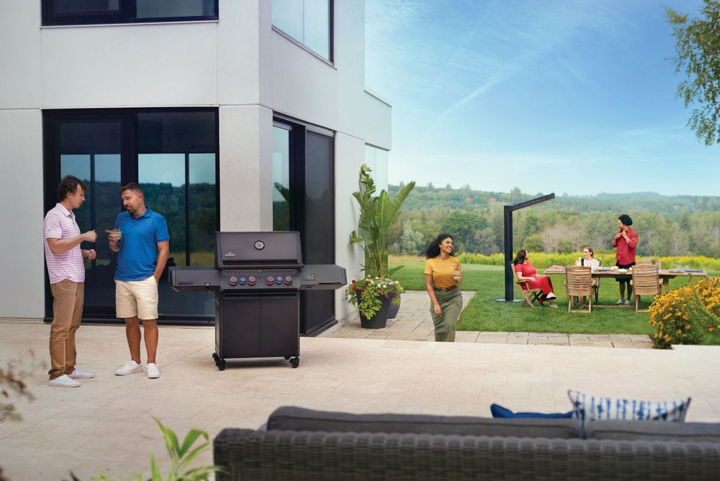 Outdoor social gathering on a patio with a grill, a modern home, plants, and people enjoying food and drinks in a scenic yard setting.