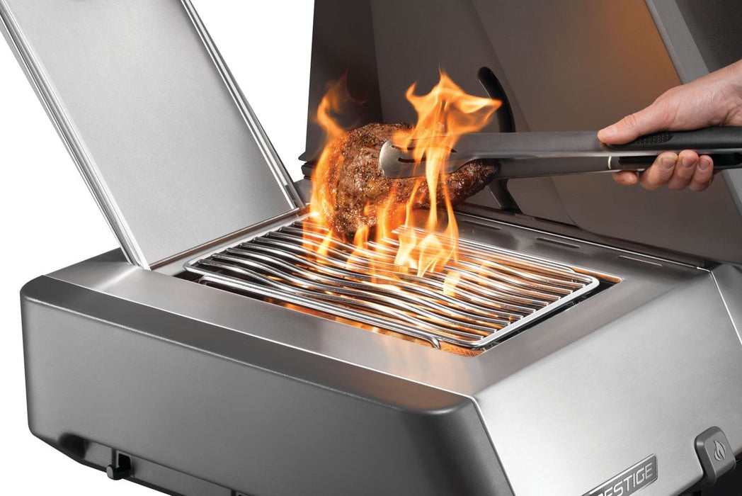 Side burner with open flame grilling a piece of meat, tongs being used to handle the food, surrounded by a stainless steel finish.