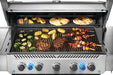 Stainless steel grill loaded with grilled vegetables, corn, steaks, skewers, and flatbread on a warming rack and main grates.