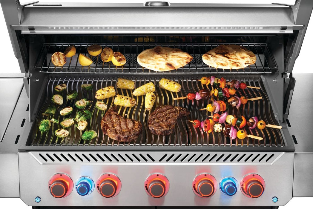 Stainless steel grill with glowing red and blue knobs, cooking vegetables, steaks, skewers, corn, and flatbreads on multiple racks.