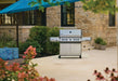 Stainless steel grill on wheels placed in a landscaped patio with stone walls, greenery, flowers, and blue umbrella-covered seating area.