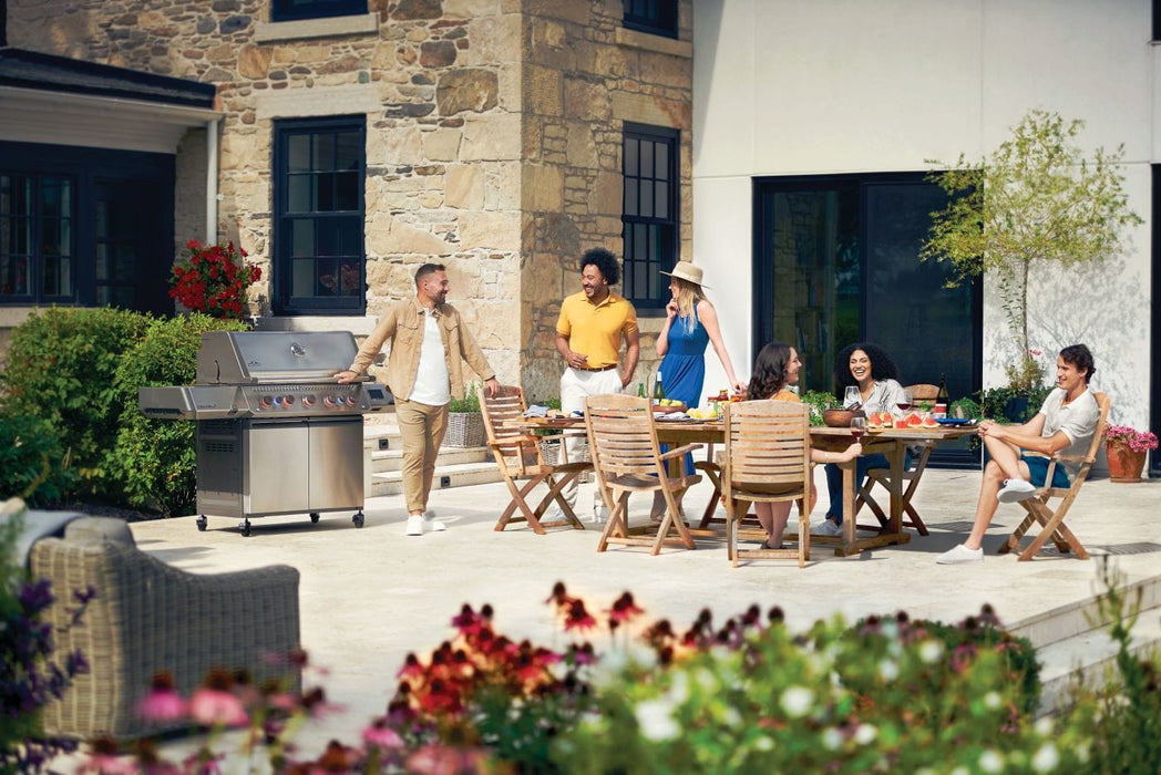 Outdoor gathering with six people enjoying a patio setting featuring a grill, wooden dining table, chairs, and vibrant garden flowers.