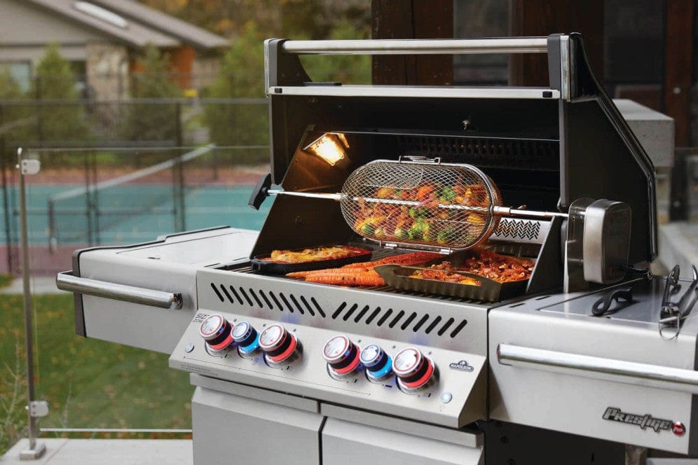 Stainless steel gas grill with open hood, interior light, skewers cooking, trays of vegetables, and meats, set on a patio with tennis courts in background