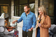 A man grilling on a stainless steel gas grill while a woman watches with a glass of wine, set on an outdoor patio