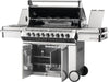 Open view of stainless steel gas grill with open hood showing rotisserie setup, side burners, control knobs, and open cabinet storage below