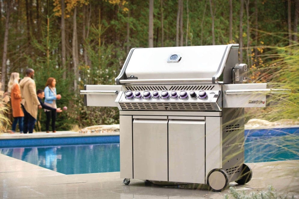 Close up view of stainless steel gas grill on a patio by a pool, surrounded by trees and people socializing in the background during autumn