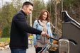 Man grilling vegetables and meat on an outdoor grill while a woman watches, set in a scenic backyard with trees in the background