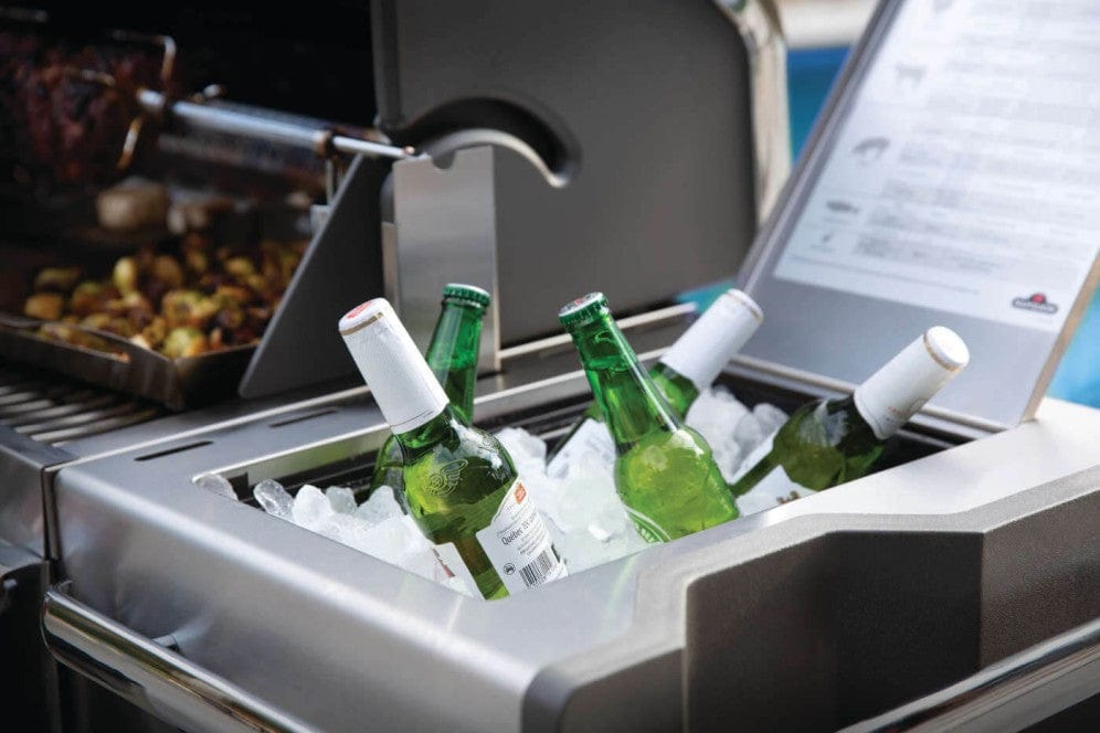 Close-up of an integrated ice bucket on a grill side shelf, chilling green beer bottles with cooking food visible in the background