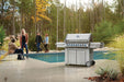 Outdoor stainless steel gas grill on a patio by a pool, surrounded by people socializing in a scenic backyard with autumn trees