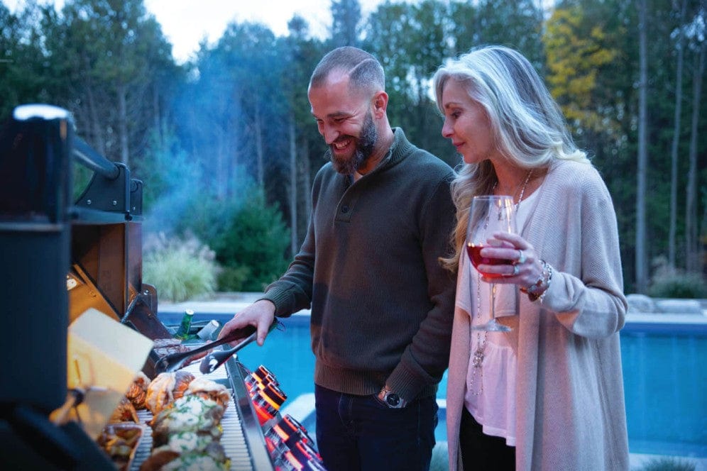 A couple grilling food by a poolside in a backyard setting, enjoying the evening with one holding a glass of wine and smiling