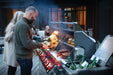 A person grilling steaks, vegetables, and corn on a lit outdoor grill, with drinks chilling in the side compartment and guests in the background