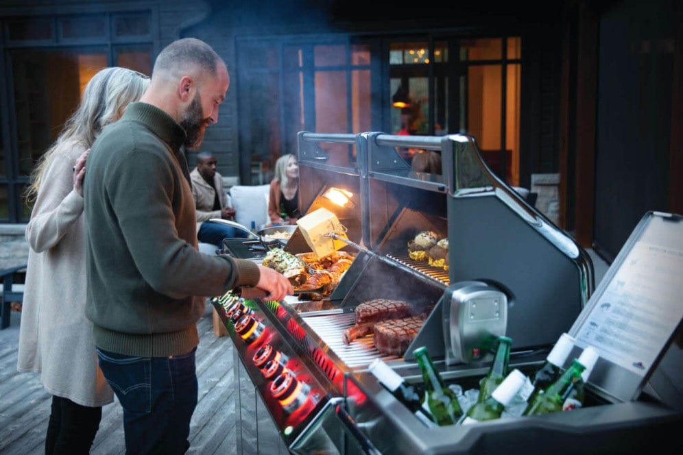 A person grilling steaks, vegetables, and corn on a lit outdoor grill, with drinks chilling in the side compartment and guests in the background