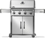 Silver freestanding outdoor grill with four control knobs, a thermometer on the lid, foldable side shelves, and double-door storage below.