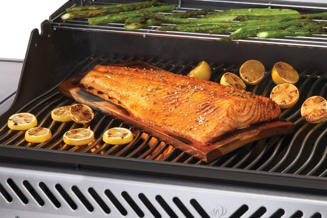 Grilled salmon on a cedar plank with charred lemon slices on grates, accompanied by asparagus spears on an upper warming rack.