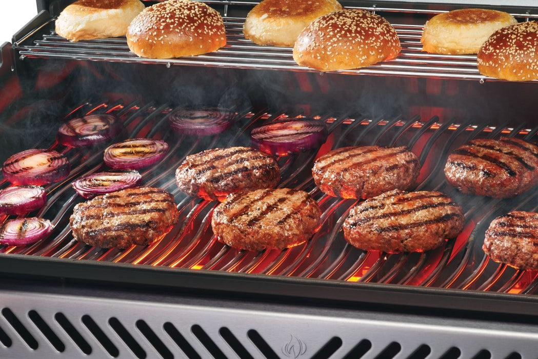 Grill loaded with sizzling burger patties and red onion slices on the main grates, with toasted buns on an upper warming rack.