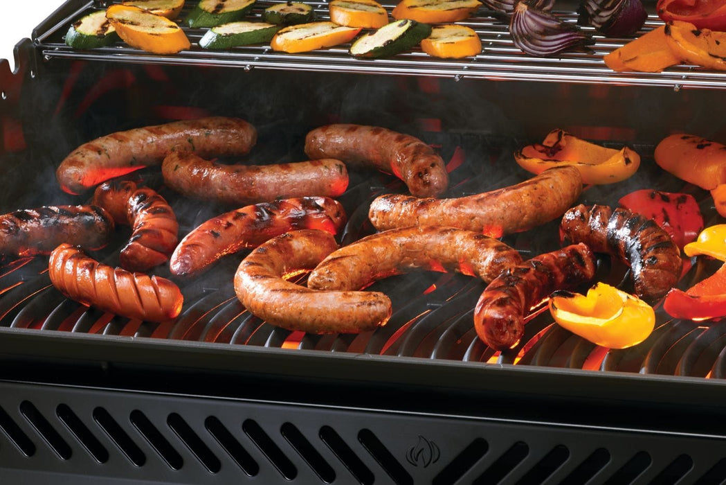 Various sausages and bell pepper slices grilling on a hot grate, with zucchini and onion slices on an upper warming rack.