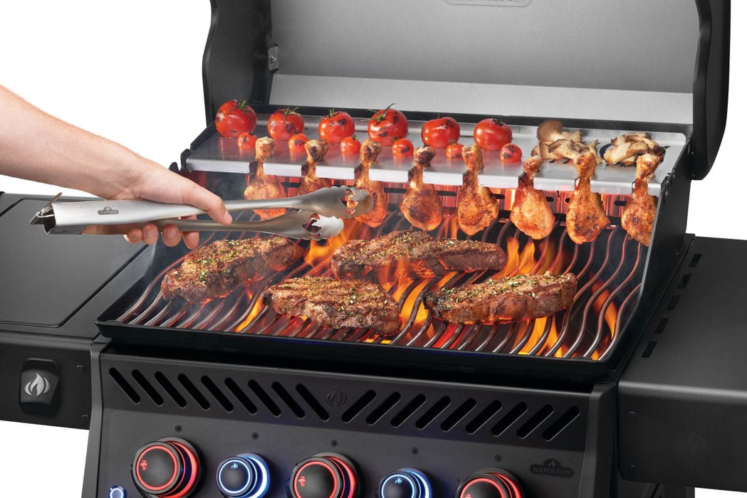 Grilling steaks and skewered vegetables on a flaming grill with a hand holding tongs, featuring vibrant controls and open hood.