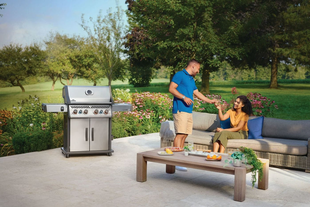 Outdoor patio scene with a couple enjoying drinks on a sofa near a barbecue grill, surrounded by lush greenery and flowers.