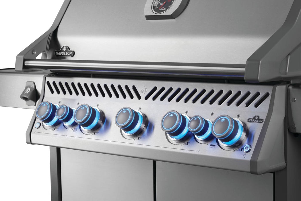 Close-up view of a grill's stainless steel control panel with six blue-lit knobs and part of the closed lid featuring a temperature gauge.