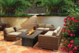 Outdoor patio setting with wicker furniture surrounding a lit fire pit, framed by vibrant flowers, palm trees, and stone stairs.