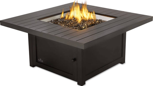 Angled view of a square fire pit with a modern metal frame, central flame, and black glass stones, ideal for outdoor spaces.