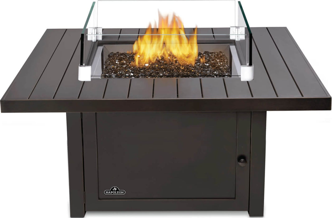 Square fire pit with black metal frame, central flame surrounded by glass panels, and decorative black stones, designed for outdoors.