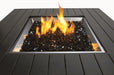 Close-up of a square fire pit featuring a black stone bed with vibrant flames, surrounded by a sleek black slatted metal frame.