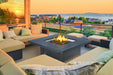 Outdoor patio with cushioned seating around a fire table, overlooking a scenic sunset view of houses, trees, and a lake.