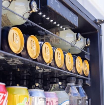 Close-up of a beverage fridge interior, showcasing neatly stacked cans, bottles, and illuminated shelves under LED lighting.