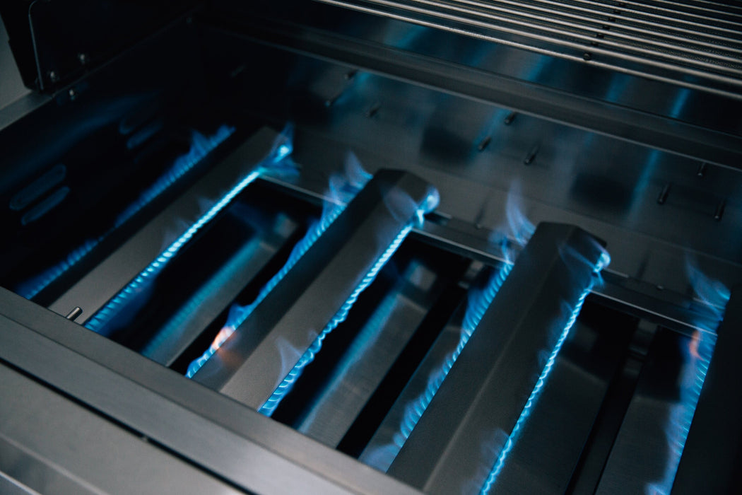 Stainless steel grill burners with blue flames lit beneath heat deflectors, showing active heating for cooking.