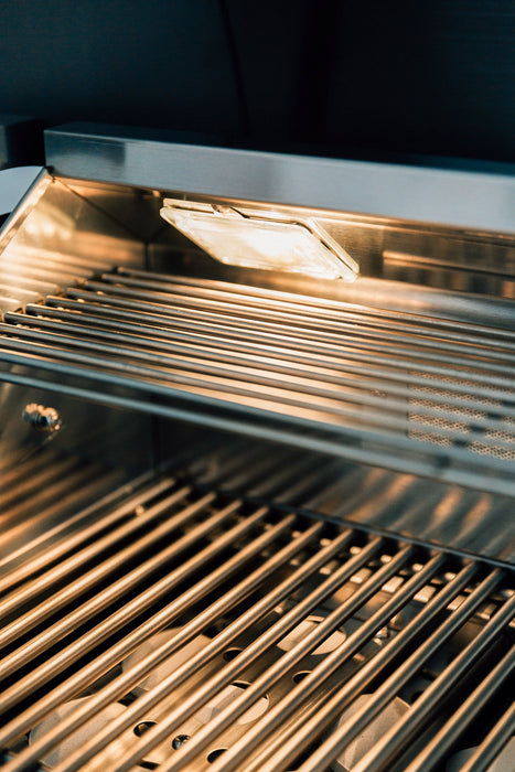 Stainless steel grill interior with lit overhead light illuminating clean grill grates and reflective surfaces.