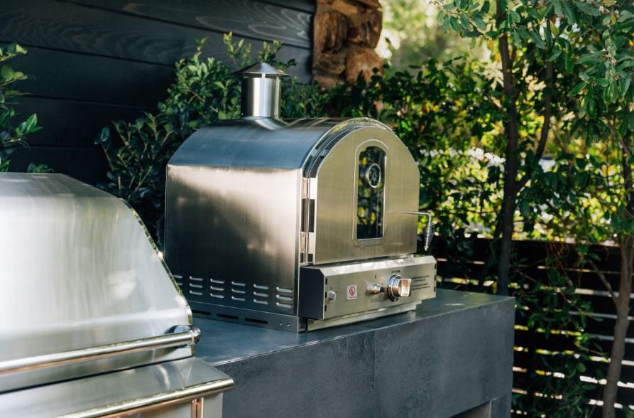 Stainless steel outdoor oven with chimney on a patio counter, surrounded by greenery and a modern black wooden wall.