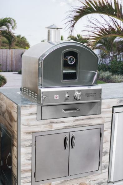 Stainless steel outdoor oven with chimney installed in a modern stone countertop, surrounded by palm trees and outdoor scenery.