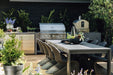 Outdoor kitchen with stainless steel appliances, pizza oven, and dining table on a wooden deck surrounded by lush greenery.