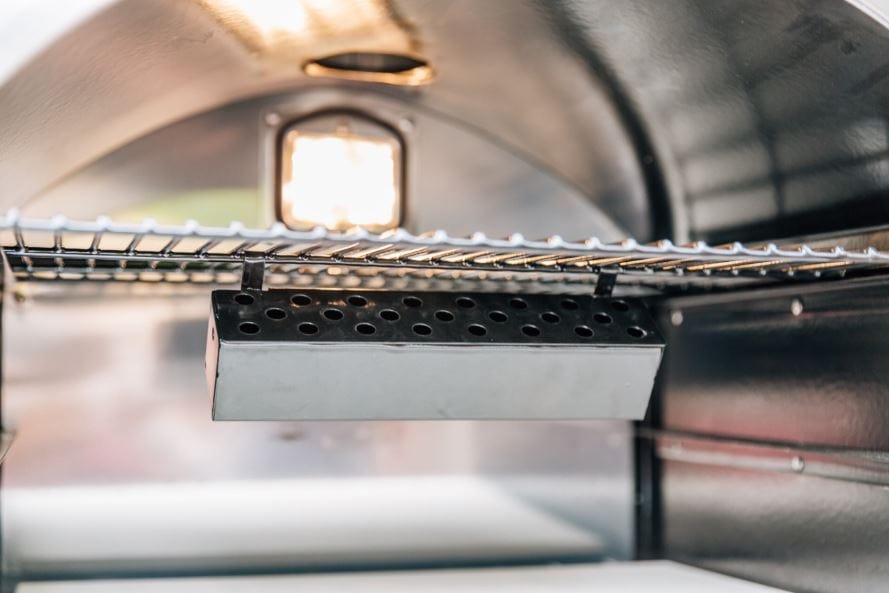 Interior of a stainless steel oven showcasing a perforated metal smoker box attached beneath a wire rack, illuminated by a built-in light.