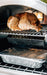 Interior of an oven with a roasted chicken on the upper rack, a smoker box beneath, and a foil-covered tray on the bottom rack.