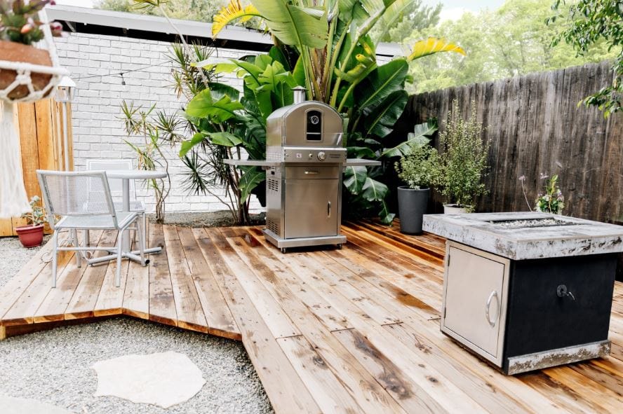 Outdoor patio setup with a stainless steel oven, lush green plants, wooden deck, metal chair, and a rustic fire pit on gravel.