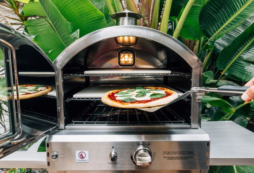 Outdoor pizza oven with a freshly baked pizza topped with basil being pulled out, surrounded by lush green tropical plants.