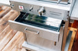 Close-up of a stainless steel outdoor oven drawer pulled out, showcasing a polished finish and wooden deck background.