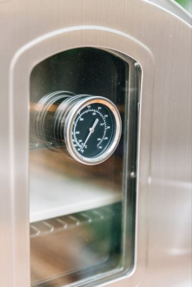 Close-up view of a stainless steel oven window with a built-in thermometer, reflecting sleek craftsmanship and interior shelves.