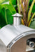 Stainless steel oven chimney with a polished finish, set against vibrant green tropical plants for an outdoor aesthetic.