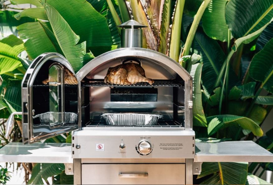 Outdoor stainless steel oven with doors open, roasting a chicken inside, surrounded by lush green tropical plants.