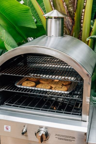 Outdoor stainless steel oven with a chimney, cookies baking on a tray inside, surrounded by tropical plants in the background.