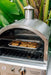 Outdoor stainless steel oven with a chimney, cookies baking on a tray inside, surrounded by tropical plants in the background.